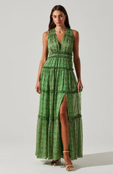 A woman is wearing a sleeveless, deep V-neck maxi dress in green with a floral pattern. The dress features tiered ruffles, an elastic waistband, and a high slit up the front, revealing her right leg. She is also wearing open-toe heels with ankle straps.